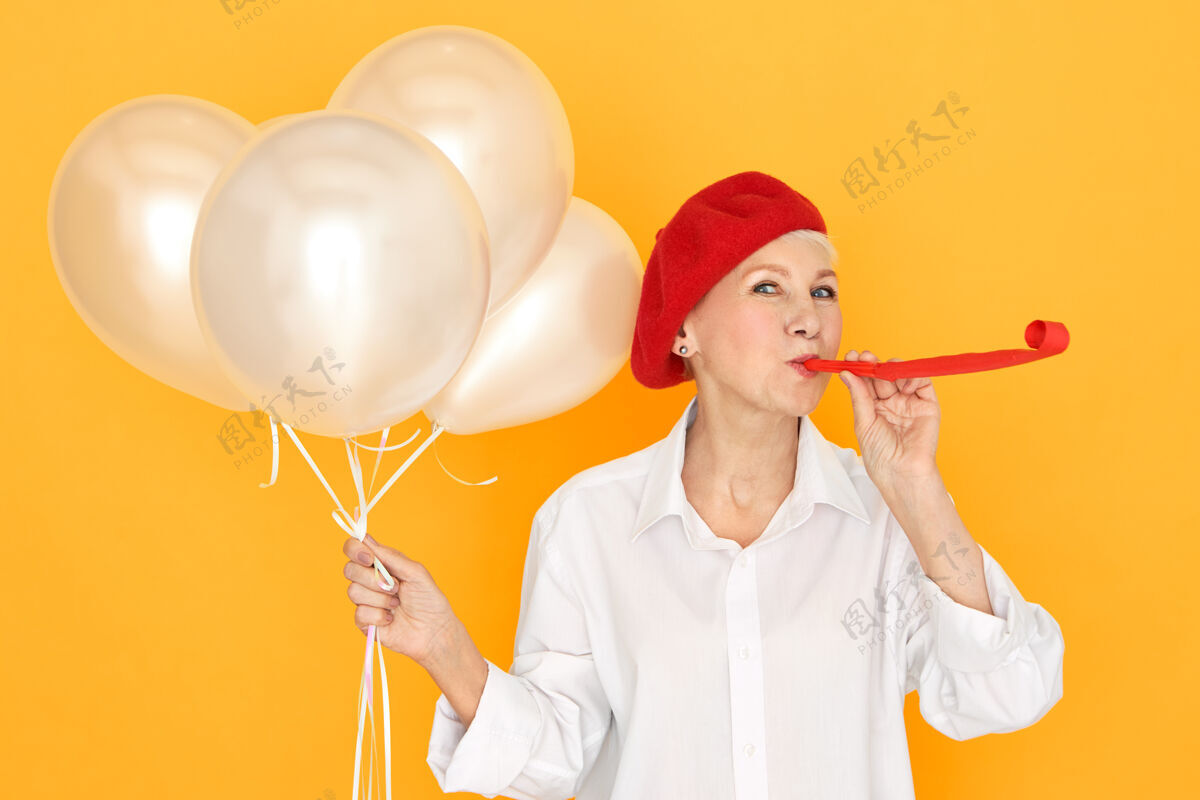 时尚一位戴着红帽子的退休妇女 享受着聚会 招待她的孙子孙女吹着口哨 手里拿着白色的氦气球生日角举行