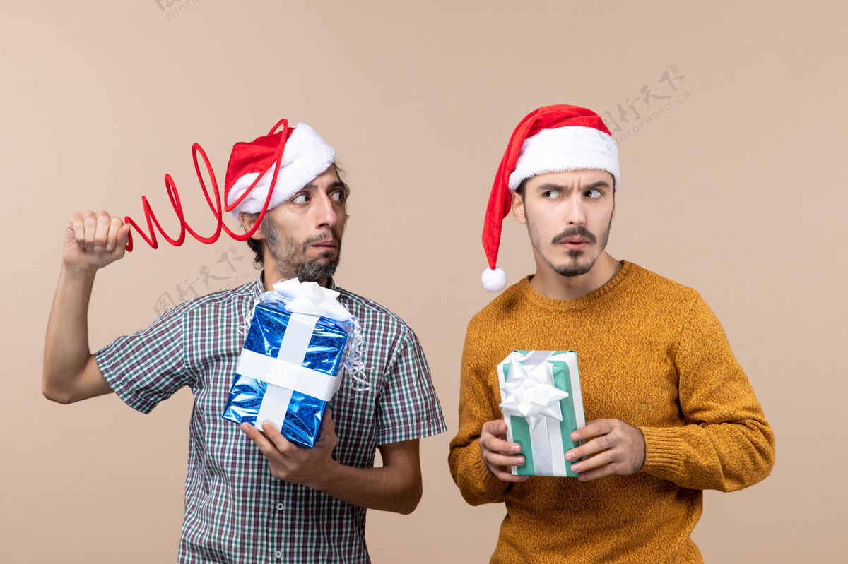 男性前视图两个困惑的家伙与圣诞帽和圣诞礼物试图集中在米色孤立的背景帽子肖像米色