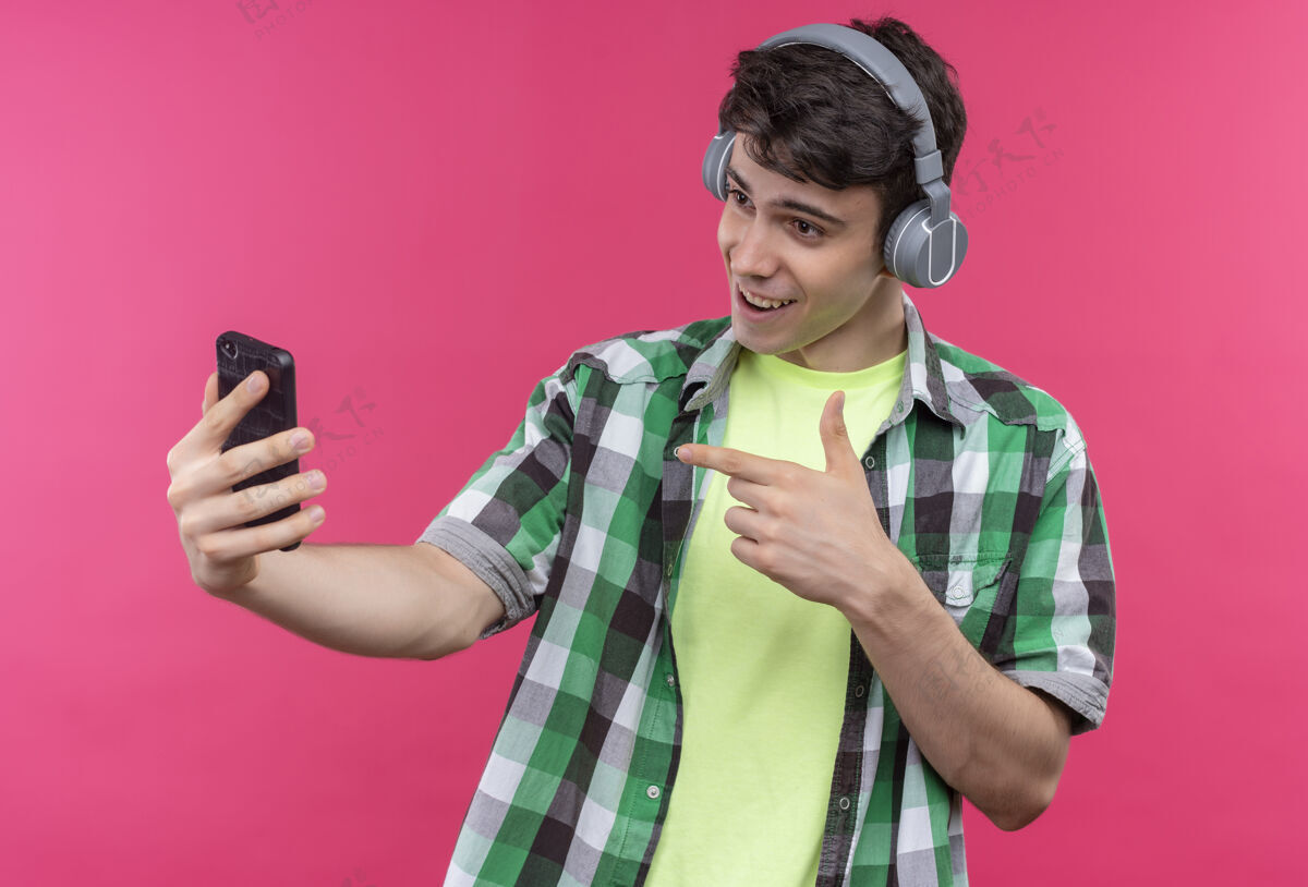 微笑微笑的白人年轻人穿着绿色衬衫 戴着耳机听音乐 手上拿着手机 背景是孤立的粉色粉色耳机绿色
