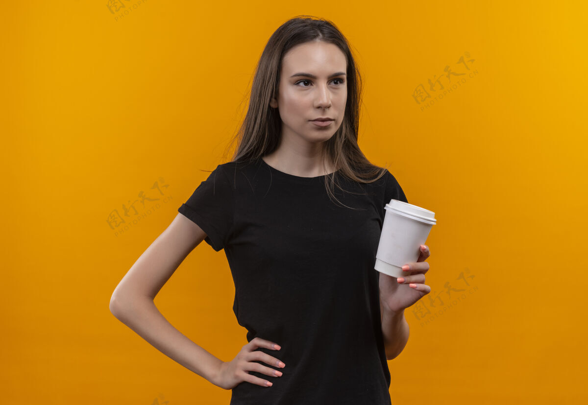 T恤看着身边年轻的白人女孩穿着黑色t恤拿着一杯咖啡把她的手放在臀部孤立的橙色背景年轻杯子拿着