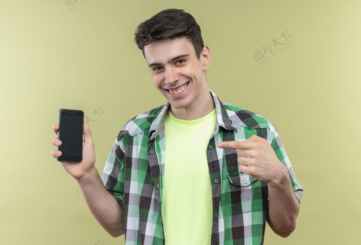 家伙一个穿着绿色衬衫的白人年轻人微笑着指着他手上的手机 背景是孤立的绿色绿色手电话