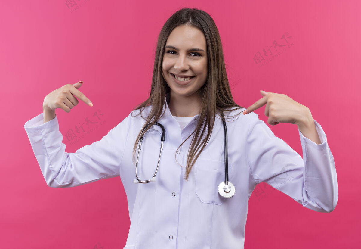 她自己身着听诊器医用长袍的年轻医生女孩微笑着指着孤立的粉红色背景上的自己微笑听诊器点