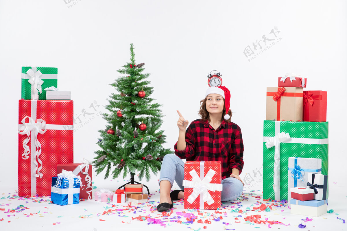 圣诞快乐前视图：年轻女子围坐在礼物旁边 头上戴着钟 站在白墙上节日礼物圣诞树