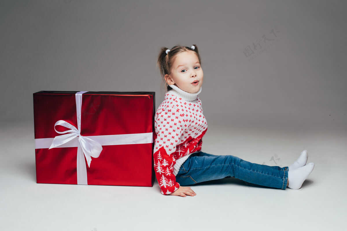 坐着可爱的小女孩 扎着马尾辫 穿着冬天的毛衣和牛仔裤 坐在红色包装的白色蝴蝶结的圣诞礼物后面孩子在镜头前撅着嘴唇侧视图圣诞快乐童年