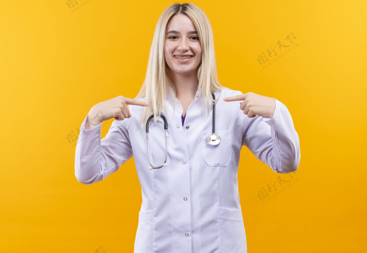 听诊器带着微笑的医生穿着医用长袍戴着牙套的听诊器的年轻女孩指着孤立的黄色背景微笑牙科点