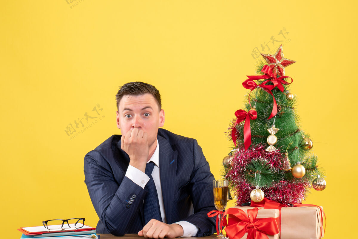 圣诞坐在圣诞树旁的桌子旁的一个好奇的男人和黄色的礼物西装董事男人