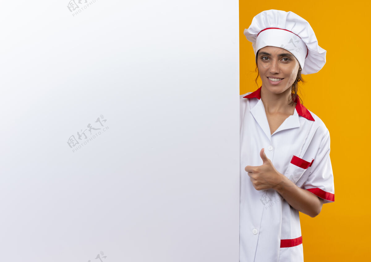 向上面带微笑的年轻女厨师身穿厨师制服 手举白墙 大拇指向上放着复印空间厨师墙女