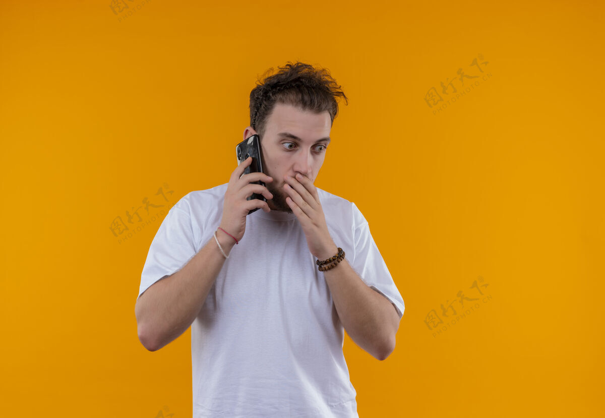 白色穿着白色t恤的年轻人在电话里讲话 手放在孤立的橙色背景上的嘴上T恤年轻嘴巴