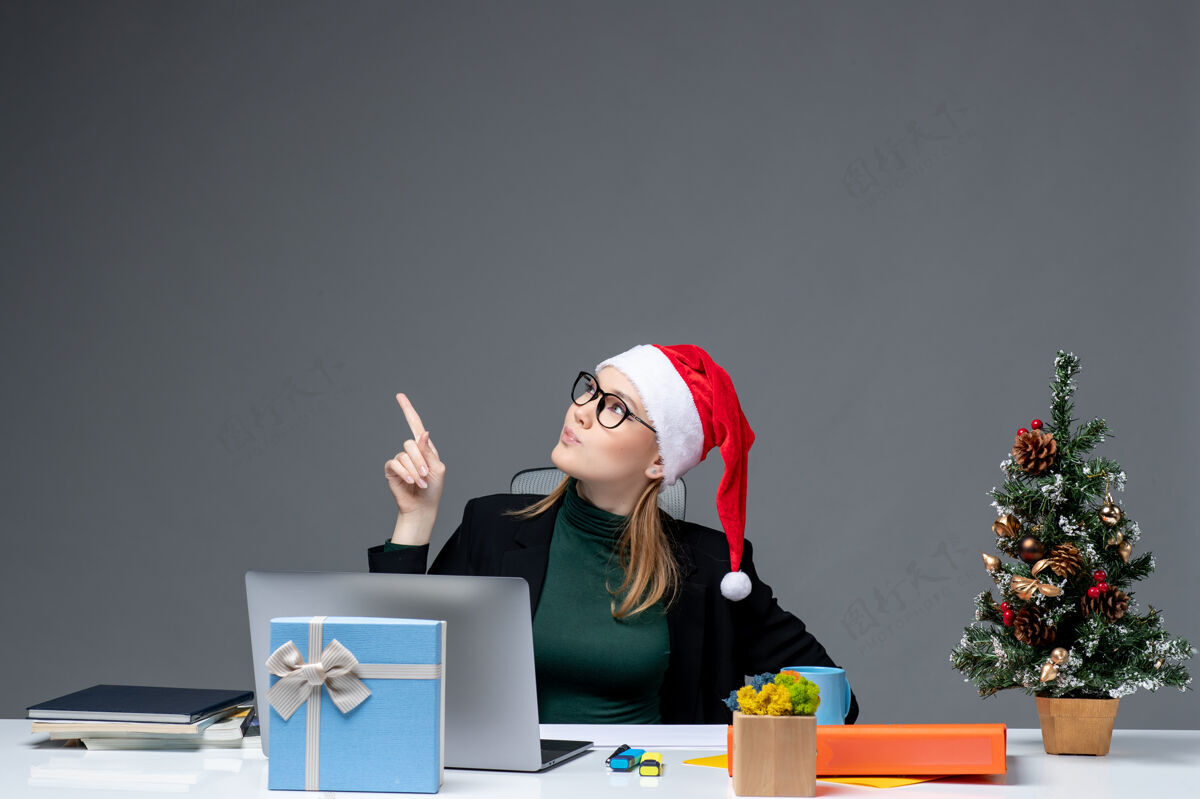 秘书一位戴着圣诞老人帽子的商务女士坐在一张桌子旁 桌子上有一棵圣诞树 还有一份礼物 在黑暗的背景下指向上方商务女士人圣诞老人