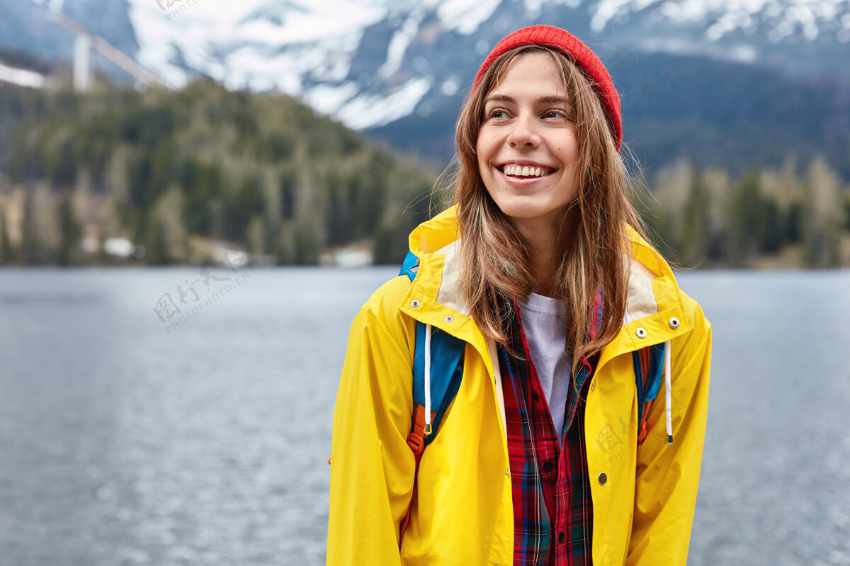 体贴年轻女游客的鲜明形象 站在山湖空间的映衬下 戴着时尚的红帽子 穿着黄色的外套背包女背包