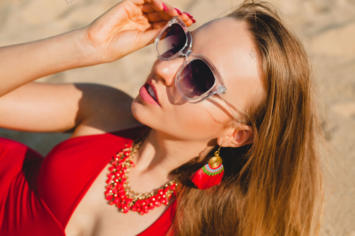 海滩年轻漂亮的金发女人穿着红色泳衣 戴着墨镜在沙滩上晒太阳耳环女人波西米亚