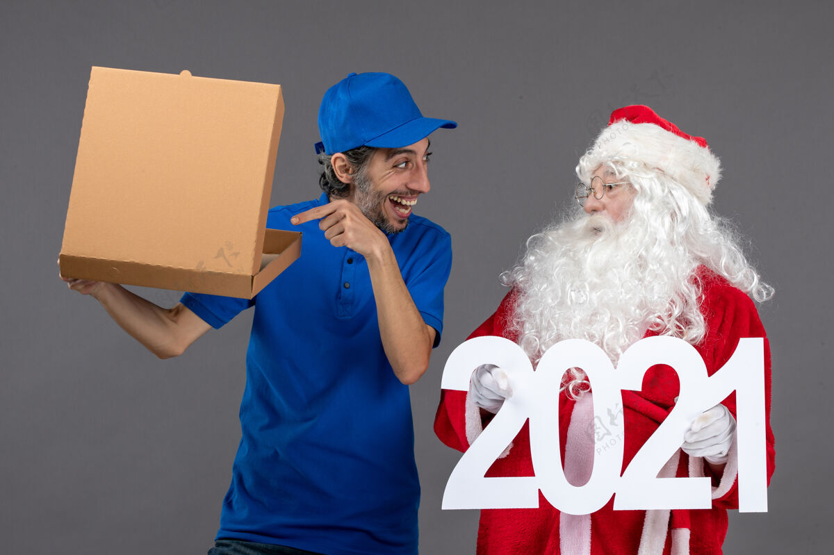 圣诞圣诞老人的正面图 男信使拿着购物袋和食品盒在灰色的墙上快乐十二月人