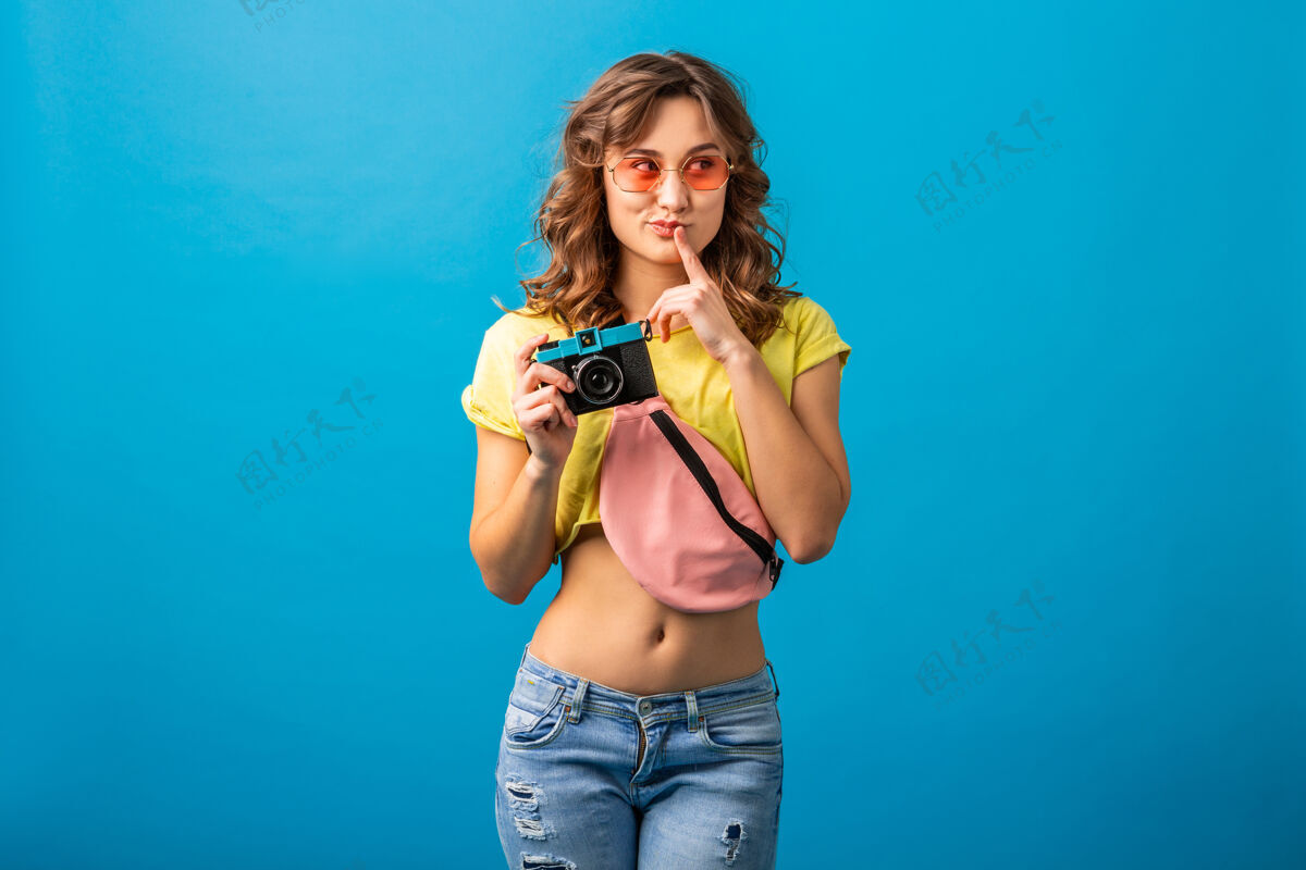 表情迷人的思考女人摆姿势与老式相机拍照穿着时髦的夏季五颜六色服装隔离在蓝色工作室的背景包脸配饰