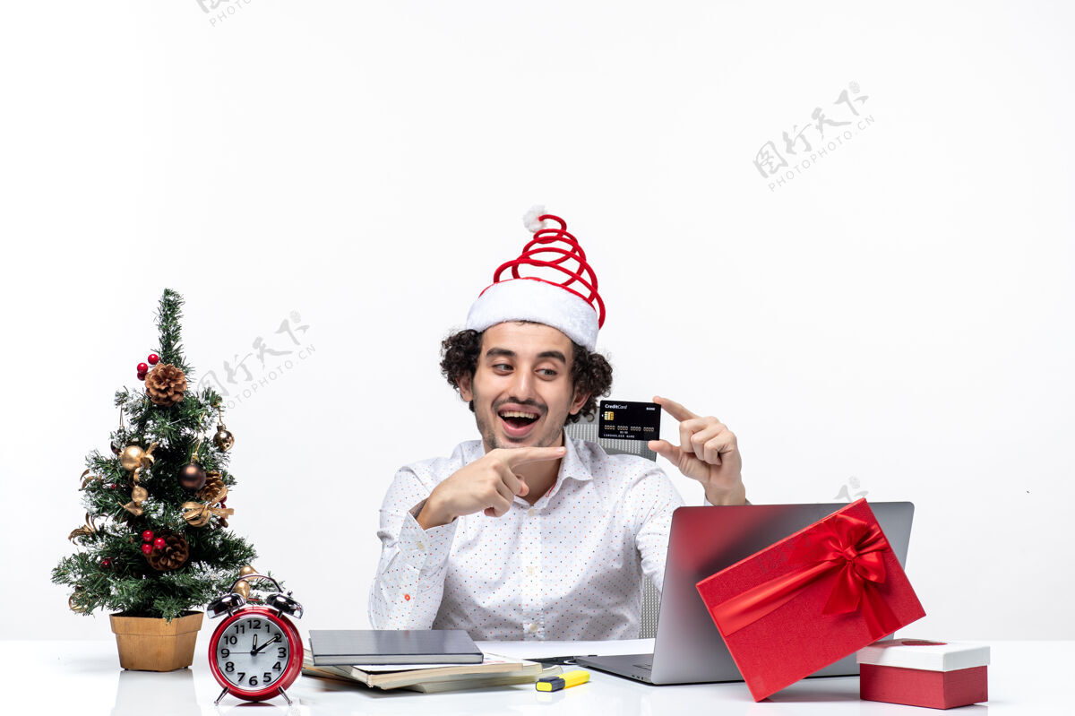 圣诞老人圣诞节心情与年轻的胡须积极的商人与圣诞老人的帽子举行 并指出白色背景上的银行卡圣诞老人指礼物