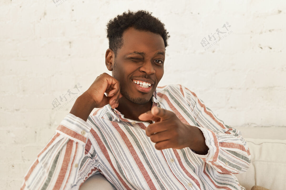 黑色室内写真帅气开朗积极向上的年轻黑皮肤男性穿着时尚衬衫坐在沙发上眨眼欢快沙发欢乐