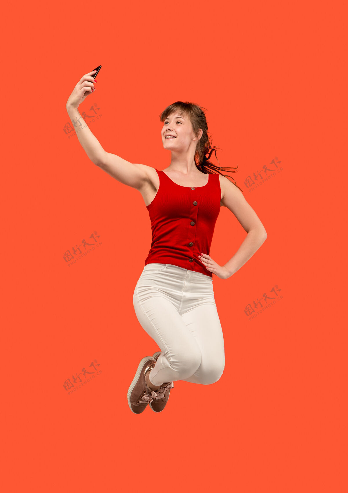 Ok一直在移动完整长度的漂亮年轻女子拿着手机和自拍 同时在红色工作室背景下跳跃移动 运动 运动 商业概念能量自拍电话