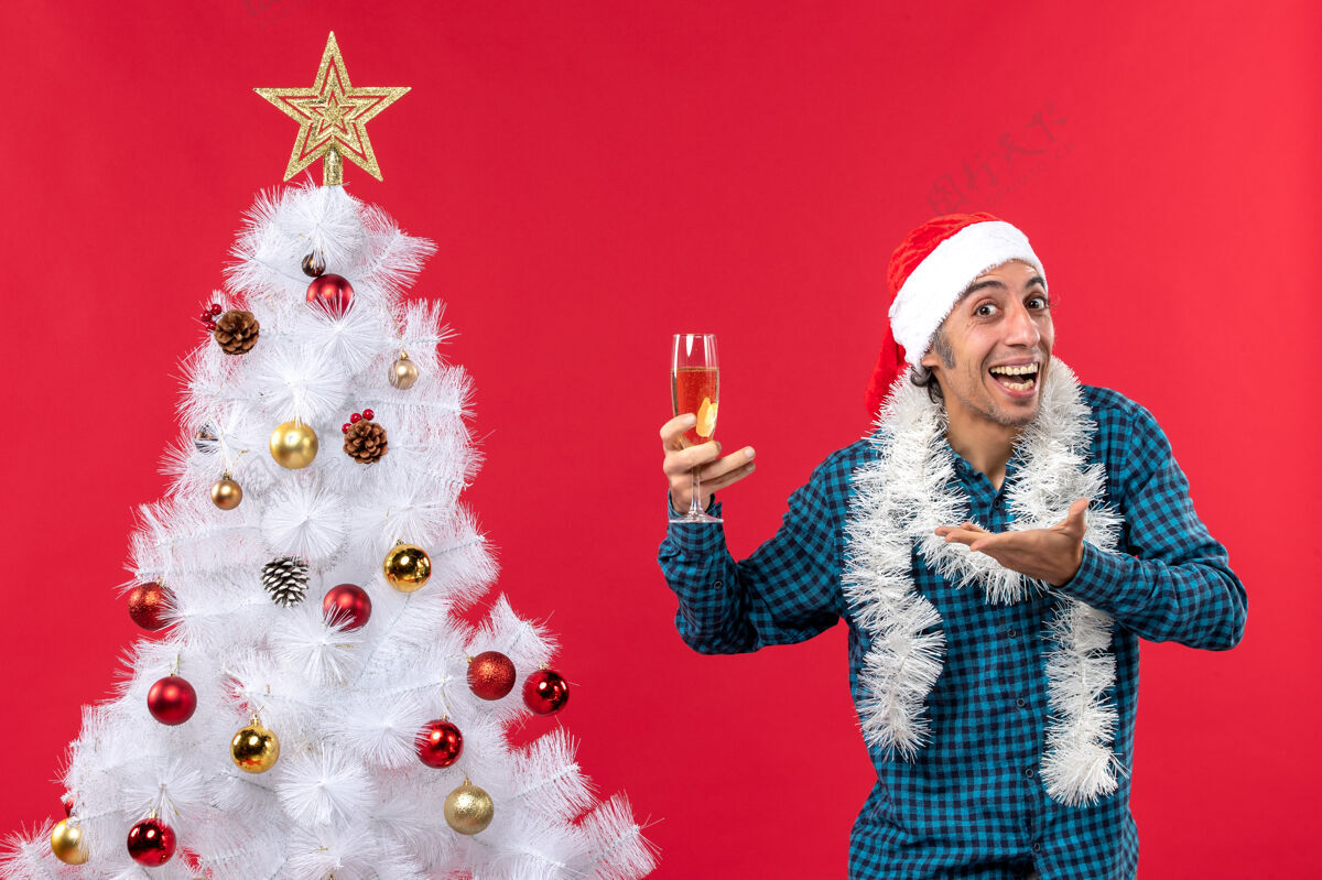 杯子圣诞节心情与情绪激动的年轻人圣诞老人帽子在一个蓝色条纹衬衫拿着一杯葡萄酒 并要求在圣诞树附近的东西问年轻人圣诞树