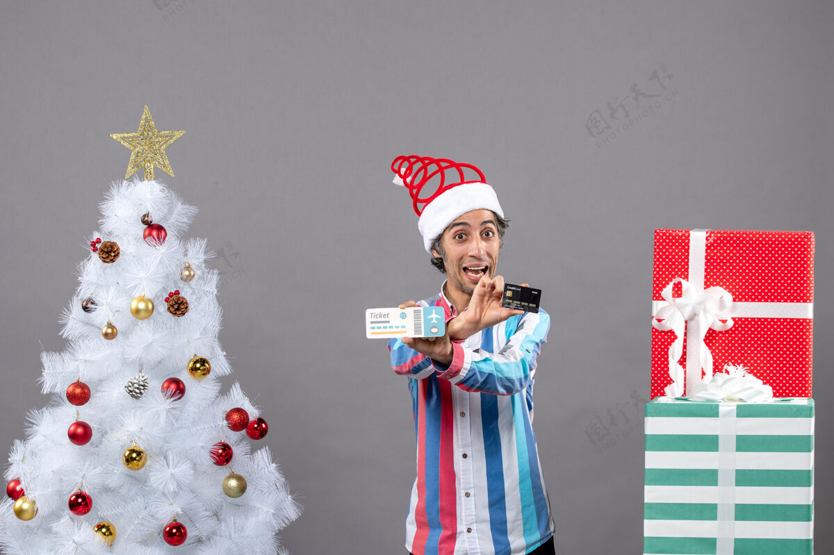 圣诞正面图：快乐的年轻人双手交叉地拿着卡片和旅行票 围着圣诞树和礼物礼物旅行年轻