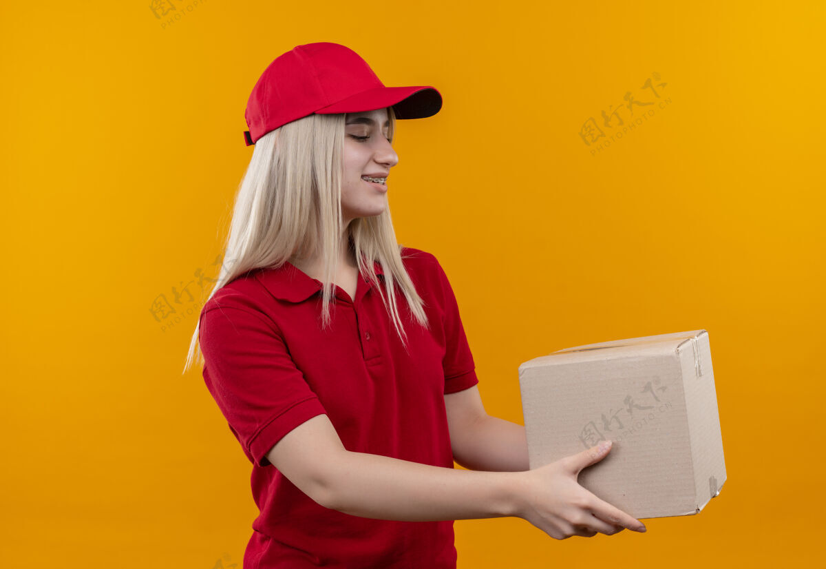 T恤微笑的小女孩 穿着红色t恤 戴着帽子 戴着牙套 旁边放着一个单独的橙色背景的盒子年轻帽子女孩