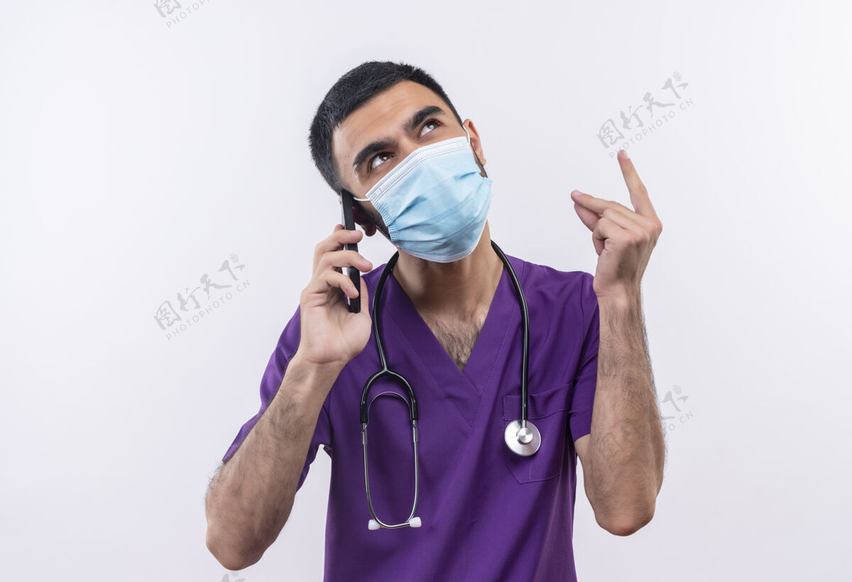男性看着身穿紫色手术服和听诊器医用面罩的年轻男医生在孤立的白色背景下讲电话戴外科医生年轻