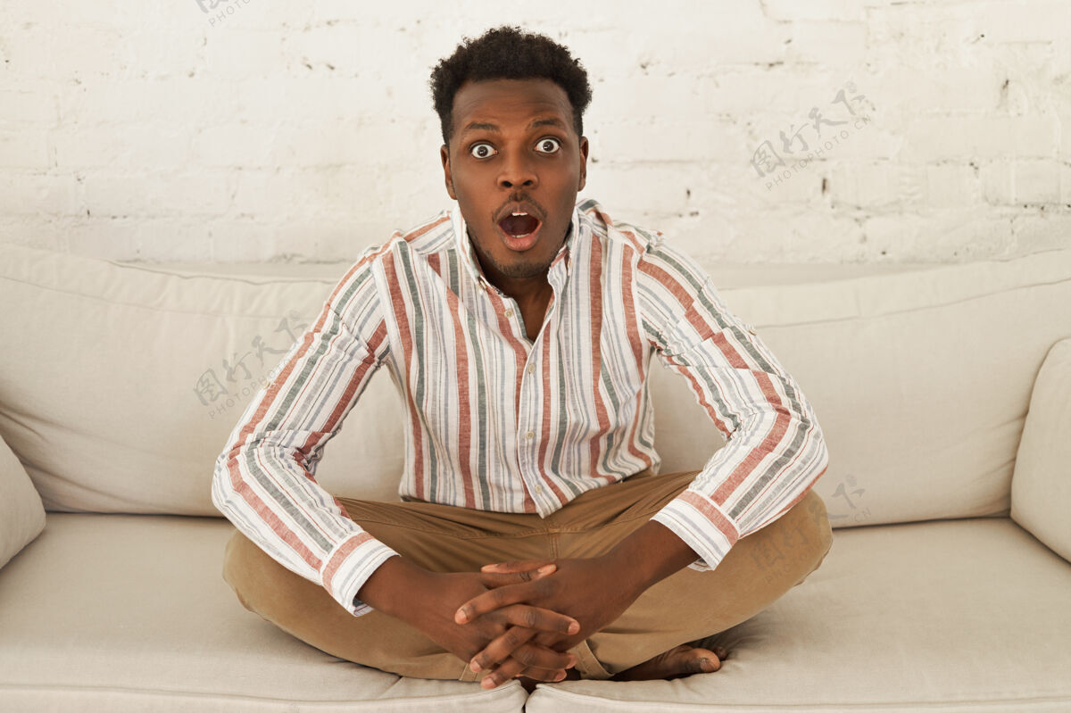 放松一个穿着休闲服的年轻非洲男人坐在沙发上 摆出一副半莲花的姿势家恐怖电视