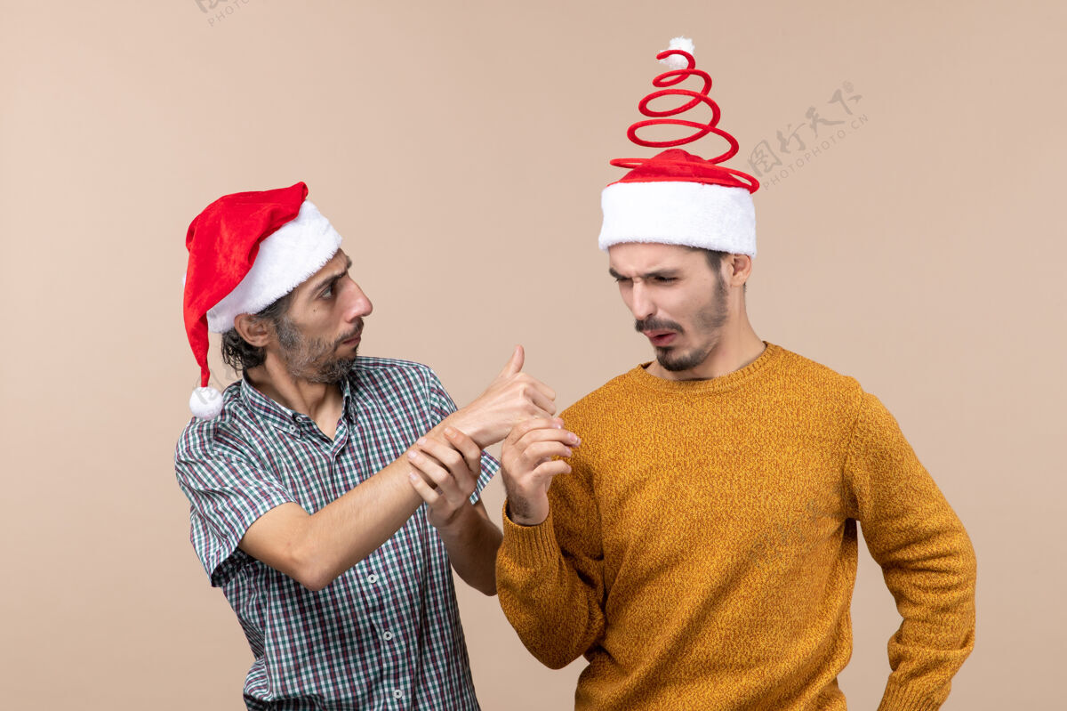 显示正面图：两个戴着圣诞帽的奇怪家伙 一个在米色孤立背景上向另一个竖起大拇指伙计们帽子前面
