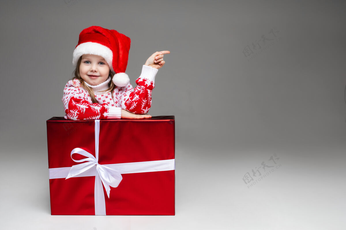 手势可爱的小女孩 穿着有图案的冬季毛衣 戴着圣诞老人帽 靠在白色蝴蝶结的包装好的圣诞礼物上可爱广告礼物