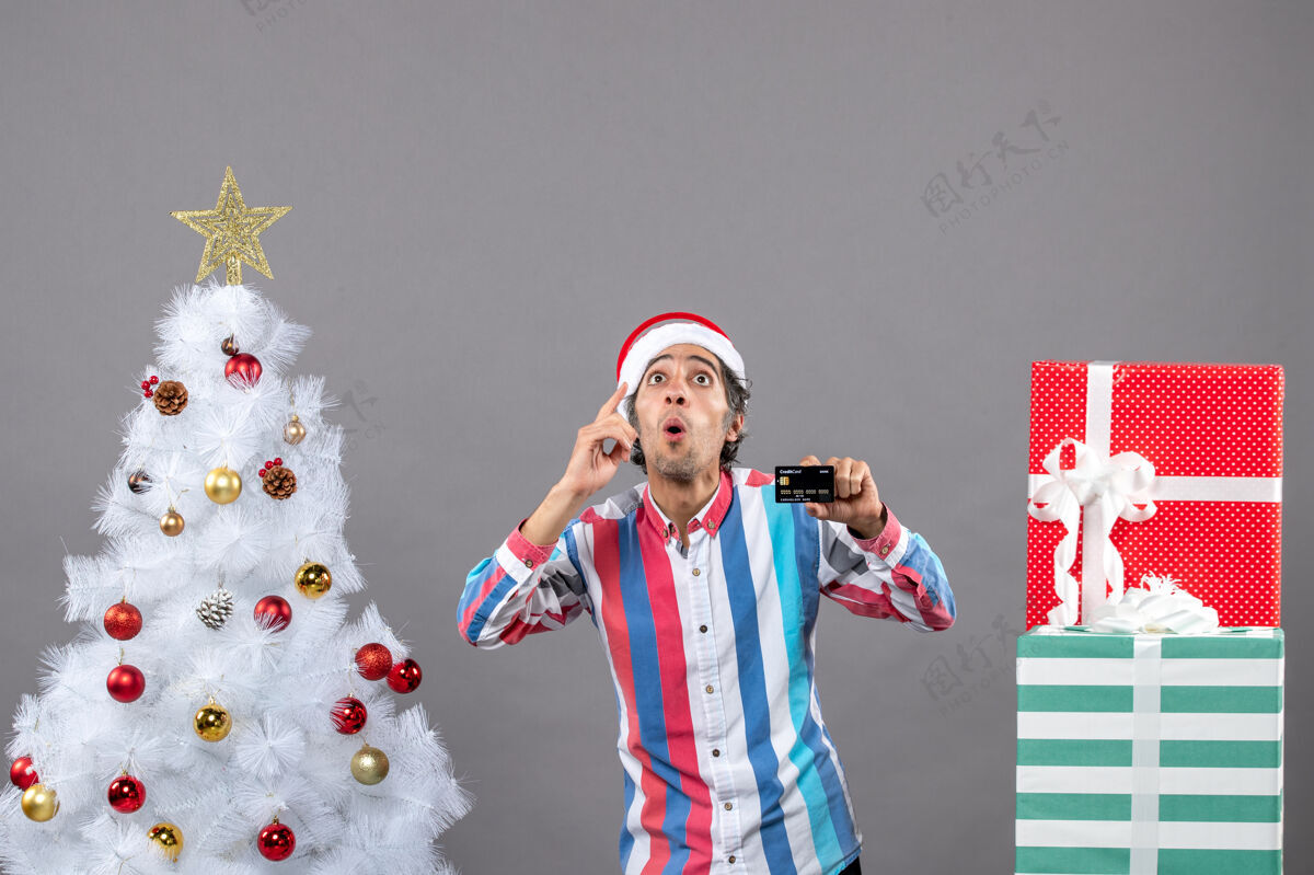 好奇的人前视图好奇的男子与信用卡站在圣诞树附近站立灰色人