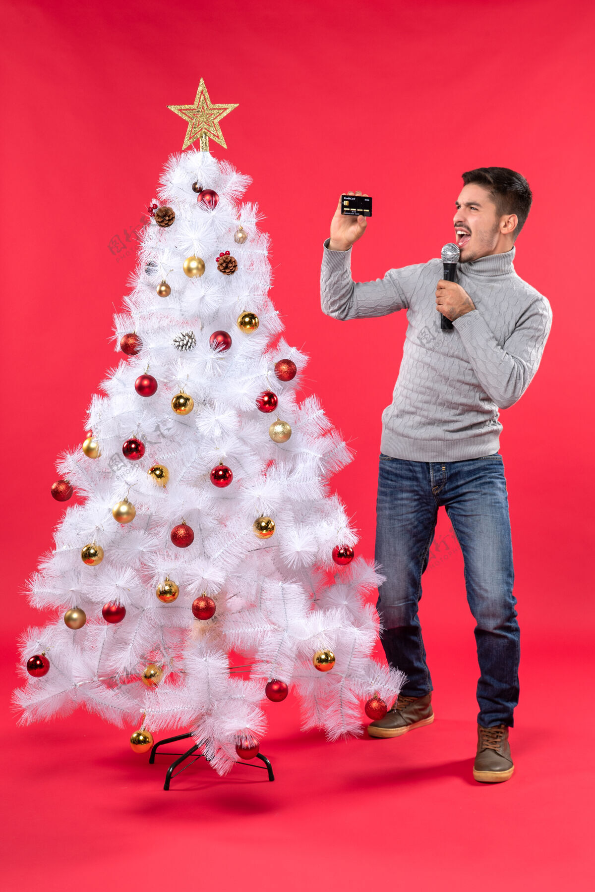 快乐圣诞节心情与自信的家伙穿着牛仔裤站在装饰圣诞树附近 手持麦克风拍摄视频自信的家伙人人