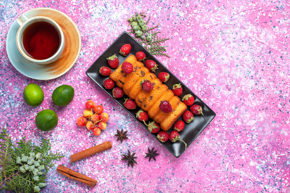 蛋糕顶视图美味的烤蛋糕放在黑色蛋糕锅里 粉红色的桌子上放着新鲜的红色草莓 茶 肉桂和柠檬烘焙草莓烘焙