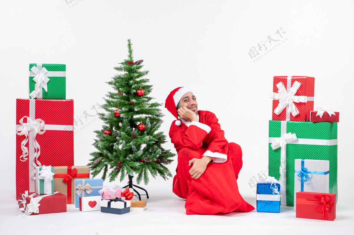 圣诞老人笑容可掬的年轻人打扮成圣诞老人 拿着礼物和装饰好的圣诞树坐在白色背景的地上地面季节礼物