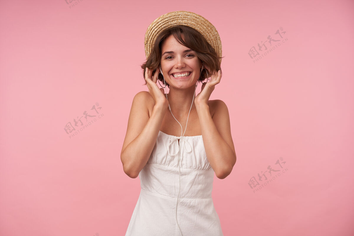 穿着快乐美丽的黑发女人 短发 穿着白色夏装 头戴草帽 戴着耳机听音乐 面带微笑 与世隔绝女性肖像表情