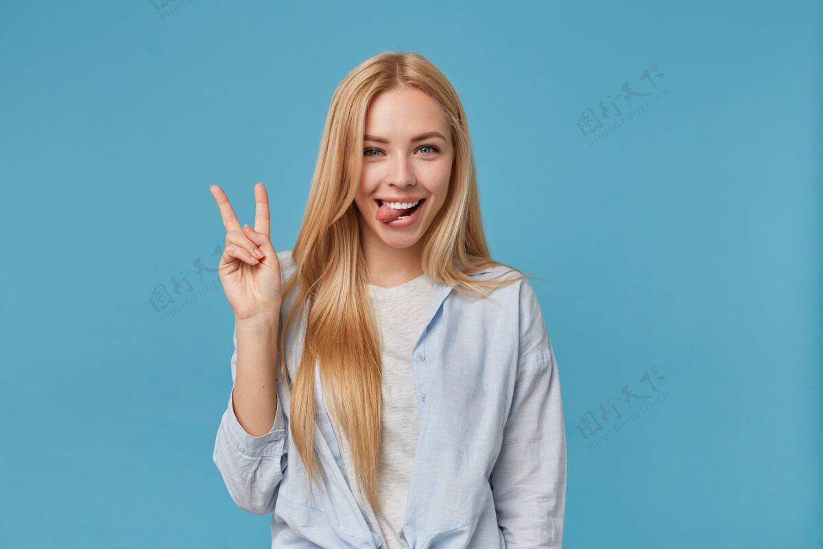 女人有趣的镜头 年轻的金发女郎 随意的发型 举起手与胜利的标志 看着喜悦的微笑 露出舌头 站在蓝色衬衫姿势金发站着