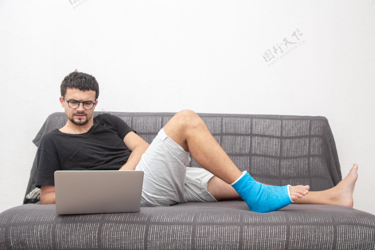 病一个戴眼镜的男人 腿骨折了 用蓝色夹板治疗脚踝扭伤 他在家沙发上用笔记本电脑工作脚恢复伤