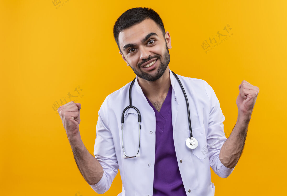 男性微笑的年轻男医生穿着听诊器医用长袍 在孤立的黄色背景上显示“是”的手势穿着礼服医学