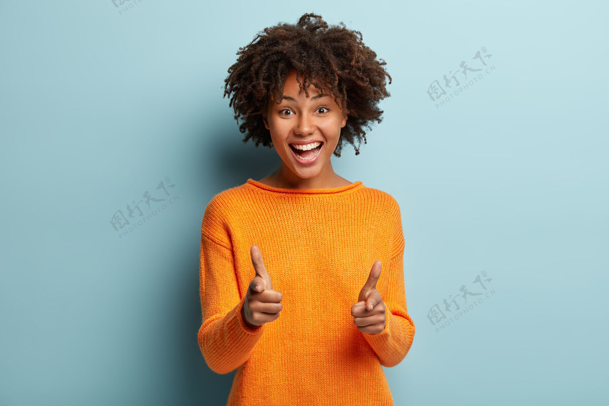 发型快乐美丽的非洲裔美国女士对着镜头做手指枪手势 表示选择 笑容灿烂 身着橙色套头衫照片发型满意