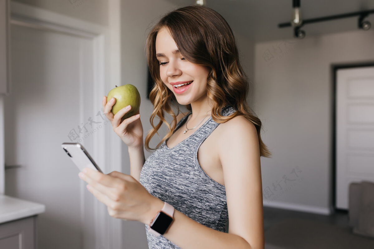 水果苗条可爱的女孩在早上使用智能手机令人惊叹的黑头发女人吃绿苹果和微笑的室内肖像有趣运动放松