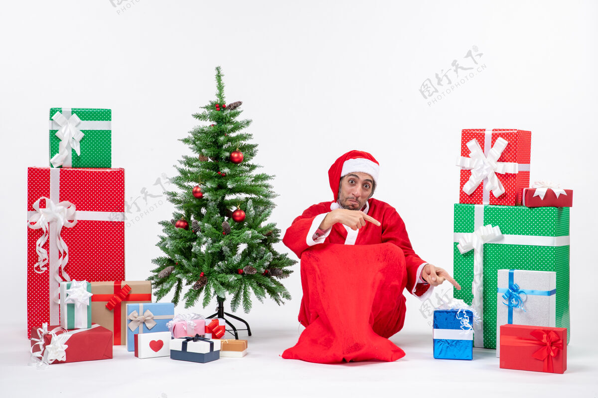 年轻人笑容满面的年轻人打扮成圣诞老人 拿着礼物和装饰好的圣诞树坐在地上指着白色的背景礼物圣诞老人圣诞树