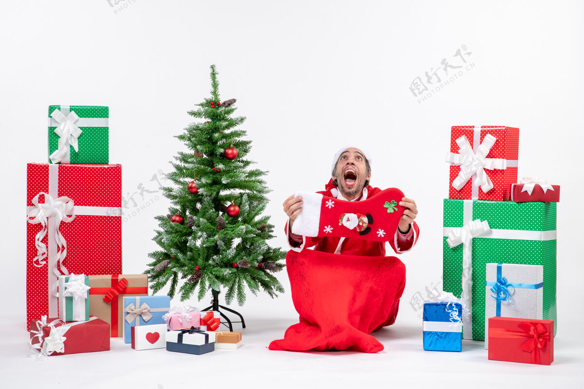 积极的圣诞老人坐在地上 在礼物旁边展示圣诞袜 白色背景上装饰圣诞树礼物圣诞树惊讶的