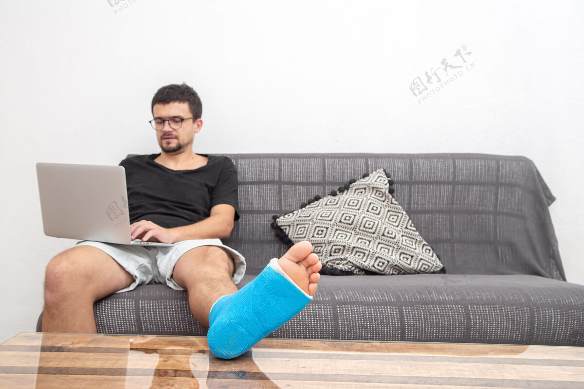 脚踝一个戴眼镜的男人 腿骨折了 用蓝色夹板治疗脚踝扭伤 他在家沙发上用笔记本电脑工作骨折伤扭伤