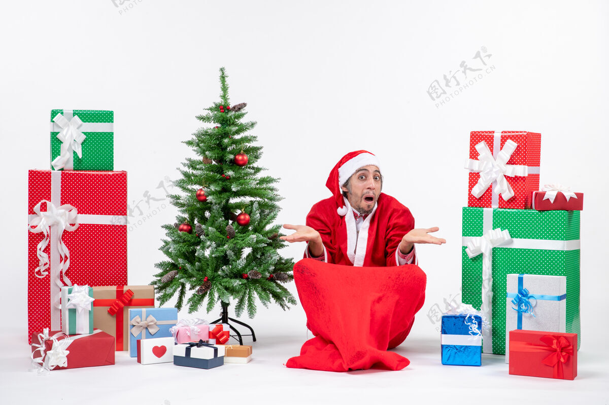 礼物惊讶的年轻人打扮成圣诞老人和礼物装饰圣诞树坐在地上的白色背景圣诞树成人人