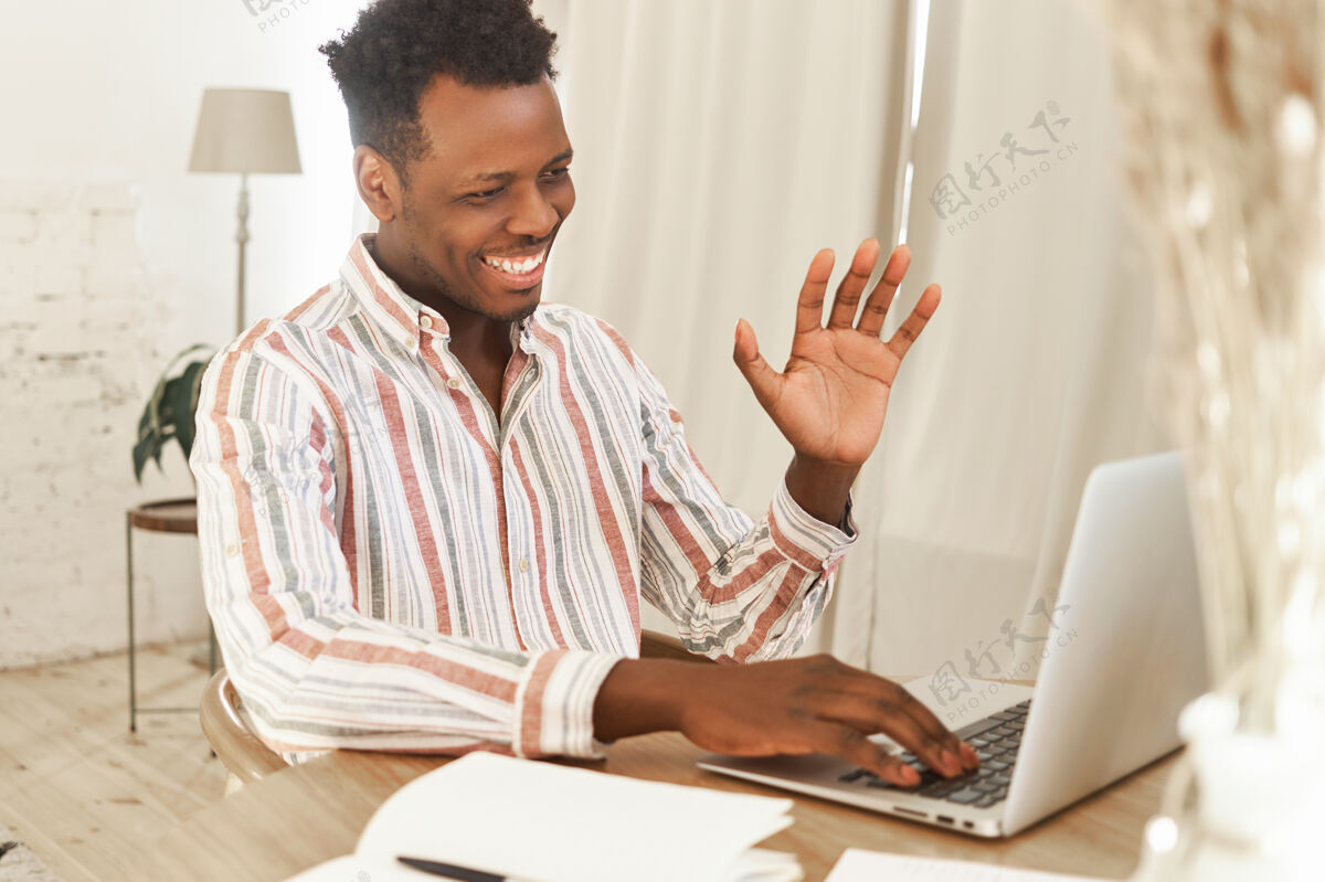 年轻开朗的非洲学生坐在打开的笔记本电脑前 面带微笑 一边用wifi在线学习 一边和导师打招呼科技海浪家