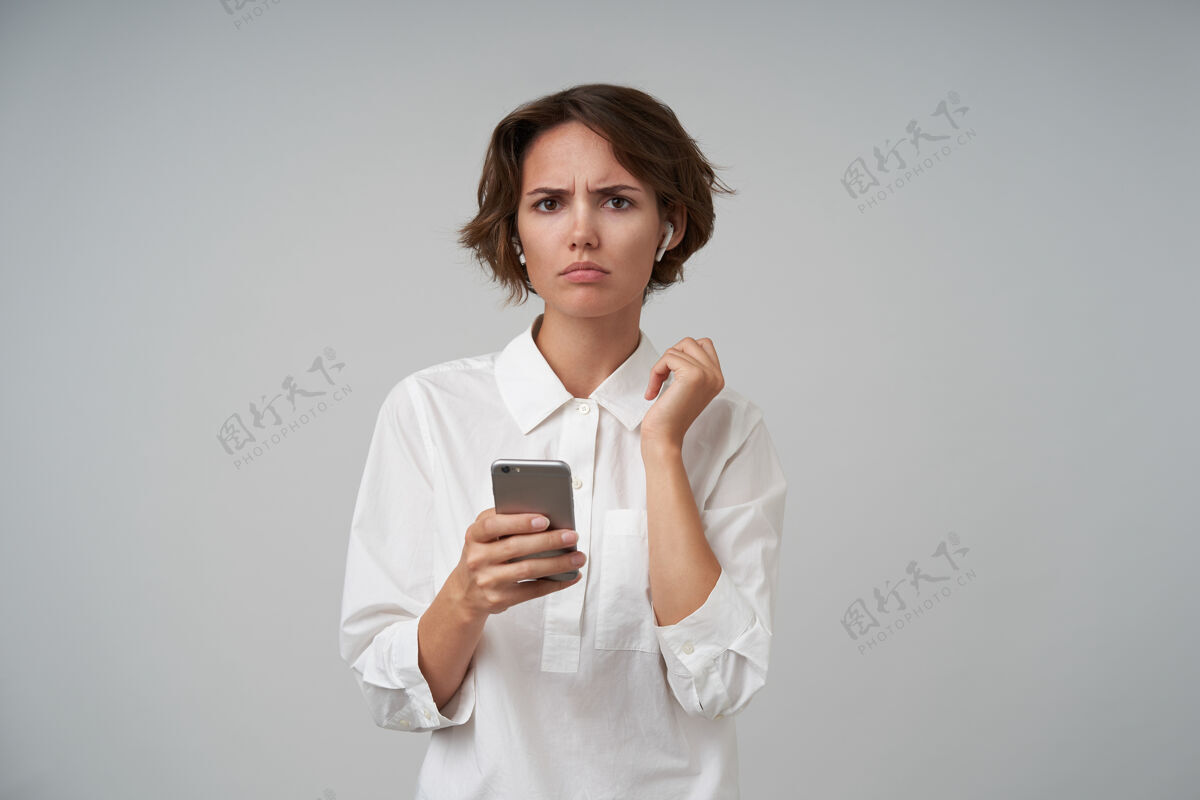 20多岁不高兴的年轻黑发女子随意的发型穿着正式的衣服 而摆姿势 眉头紧锁 手持智能手机摆姿势理发棕色