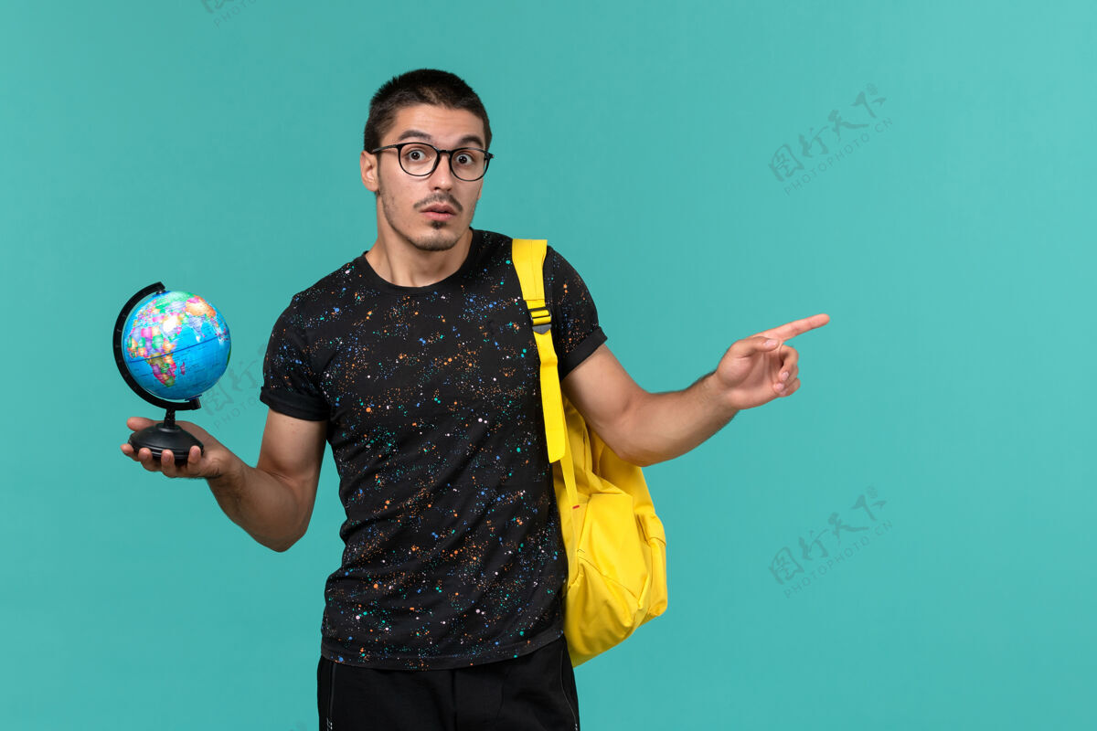人民身穿深色t恤黄色背包的男学生正面图蓝色墙上拿着小地球仪游戏游戏设备正面