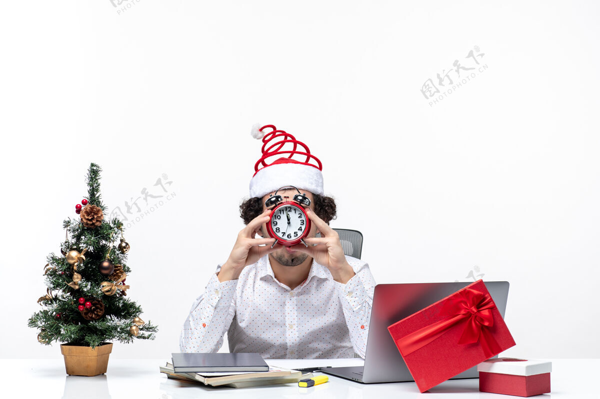 圣诞节戴着圣诞老人帽 拿着闹钟 坐在白色背景的办公室里的年轻商人圣诞老人商务圣诞老人