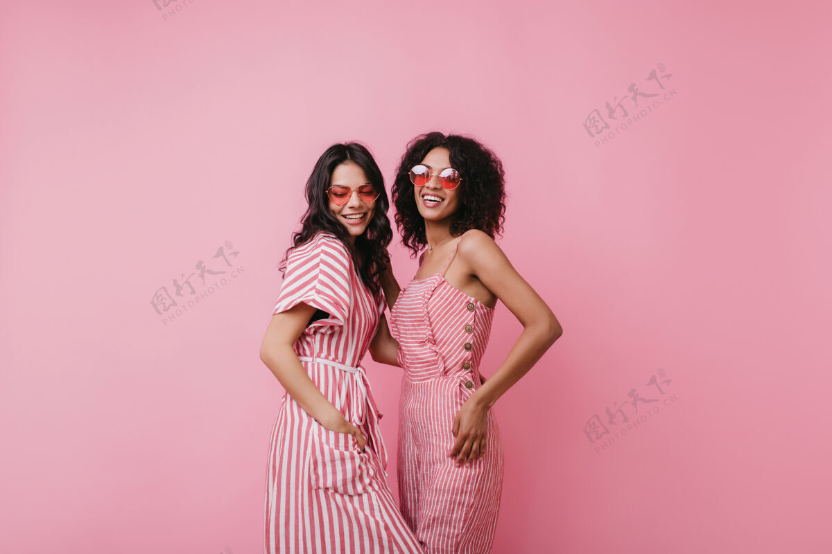 时尚身材匀称的拉丁女人穿着条纹服装和非洲女性朋友合影两位漂亮女士戴着时髦的粉色太阳镜的室内照片欢快粉色拉丁