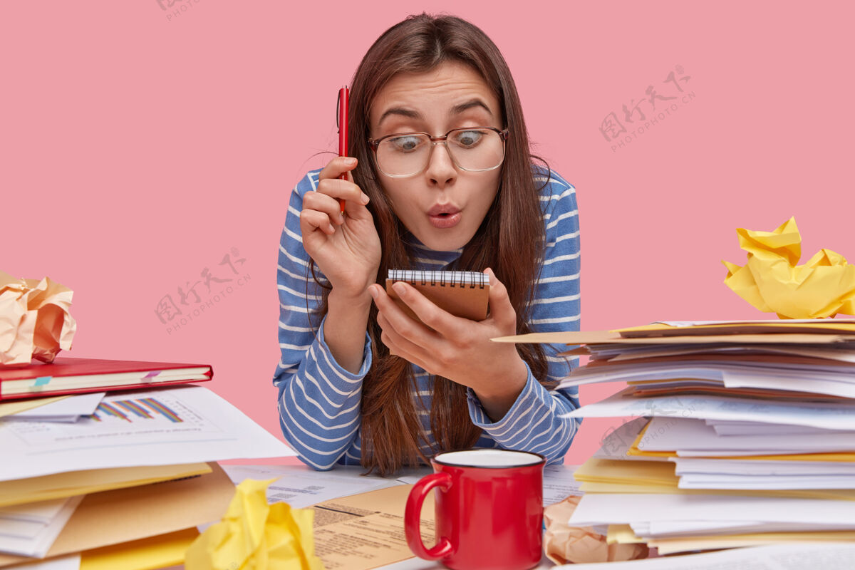饮料震惊的富尼年轻的女书生盯着笔记本 拿着笔 在工作场所有成堆的文件 戴着眼镜和条纹衣服工作区工作场所学生