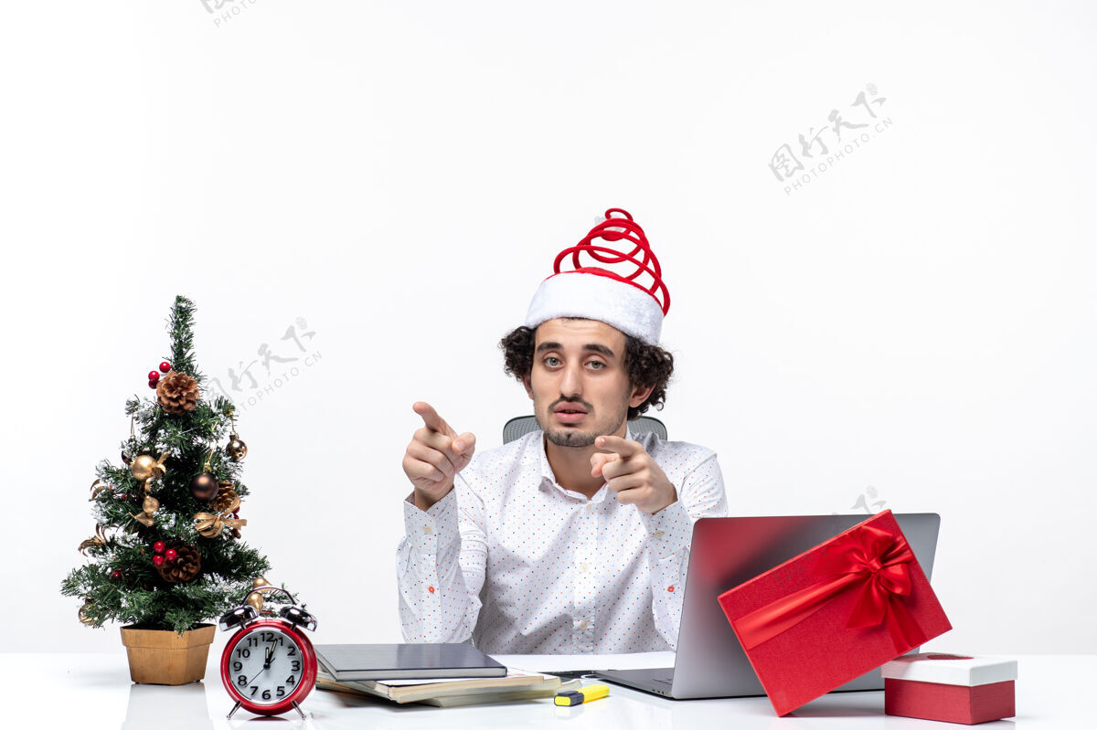 微笑困惑的年轻商人戴着滑稽的圣诞老人帽子 在白色背景上指着办公室里的东西圣诞老人年轻的商人圣诞节