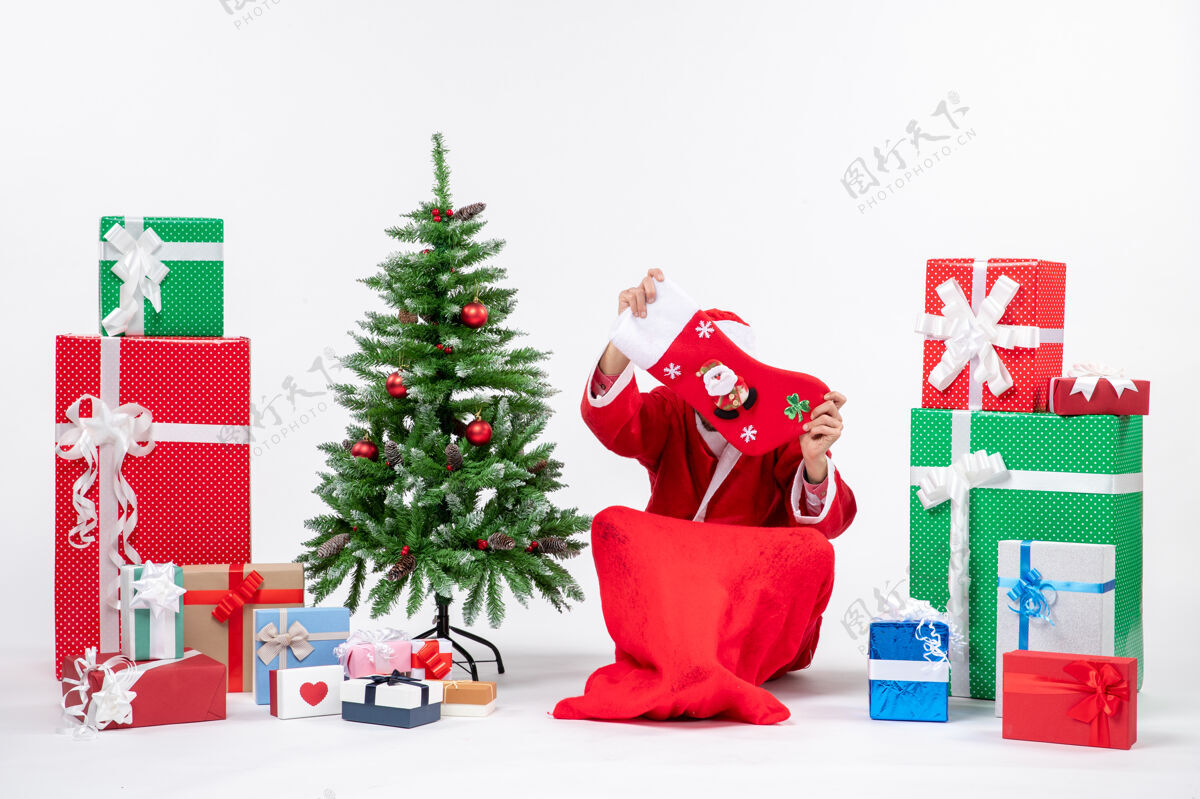 袜子圣诞老人坐在地上 在礼物和白色背景装饰圣诞树旁用圣诞袜子把脸合上 这是节日的气氛圣诞装饰结束