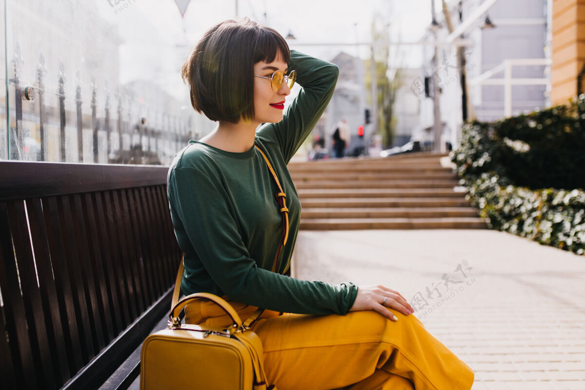 短发优雅的短发女孩戴着太阳镜在户外放松穿着绿色毛衣的可爱女人在阳光明媚的日子在长凳上摆姿势城市人户外